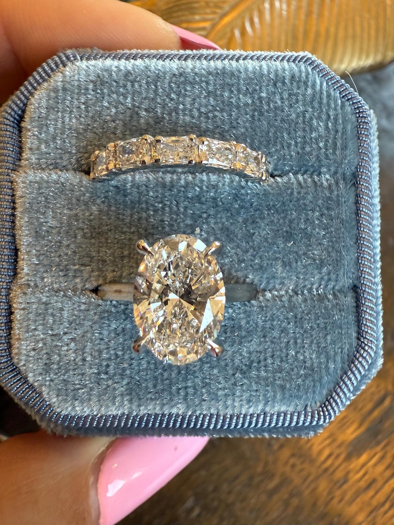 May include: Two diamond rings in a blue velvet ring box. The top ring features a band of rectangular diamonds. The bottom ring has a large oval-cut diamond set on a silver band.