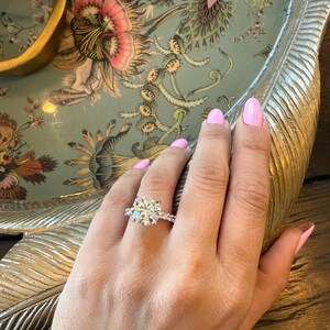 K&ouml;nnte beinhalten: Ein Diamant-Verlobungsring mit einem gro&szlig;en, rund geschliffenen Mittelstein und kleineren Seitensteinen, gefasst in einem silbernen Band. Der Ring befindet sich an einer Hand mit rosa Nagellack, vor einem floralen Tablett.