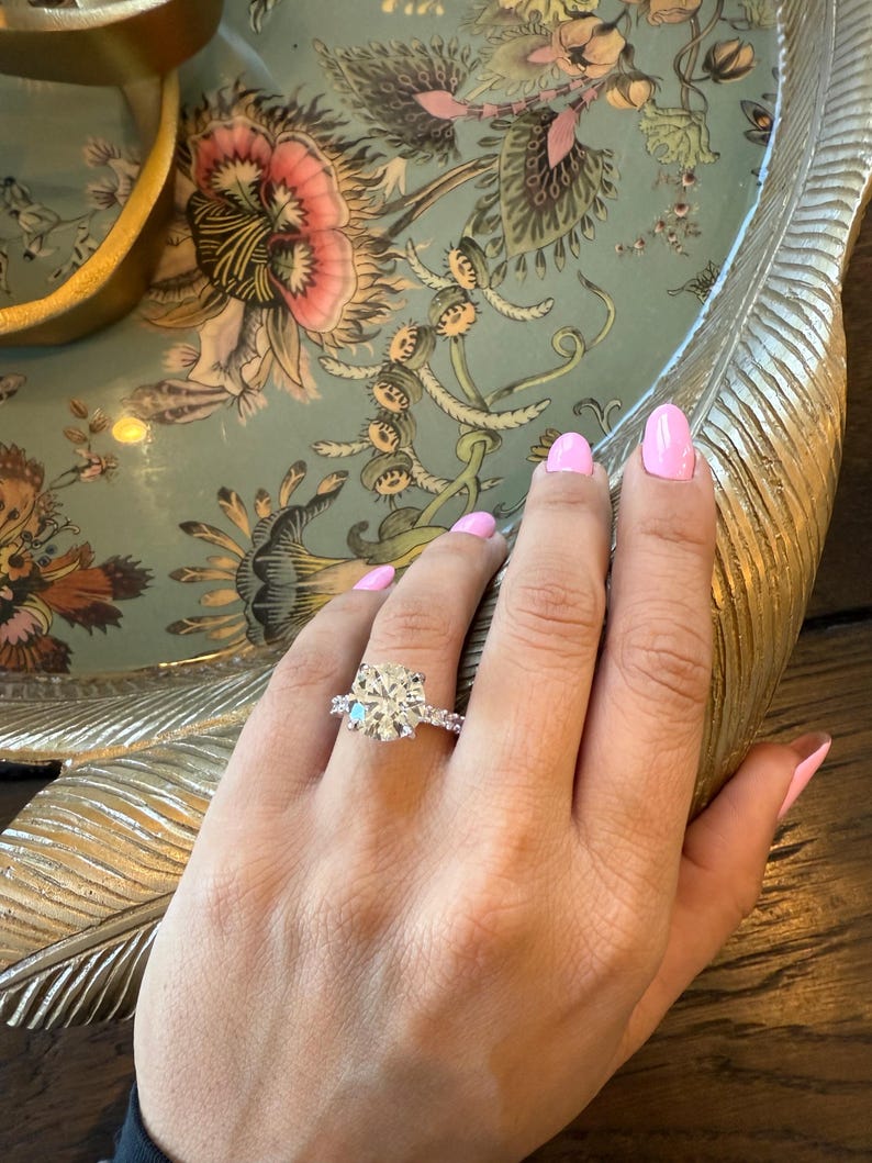 K&ouml;nnte beinhalten: Ein Diamant-Verlobungsring mit einem gro&szlig;en, rund geschliffenen Mittelstein und kleineren Seitensteinen, gefasst in einem silbernen Band. Der Ring befindet sich an einer Hand mit rosa Nagellack, vor einem floralen Tablett.