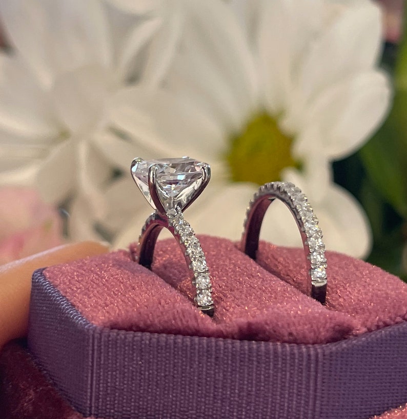 May include: A pair of engagement rings in a pink velvet ring box. The main ring features a large, clear, square-shaped diamond with a pave setting. The band of the main ring is also pave set with smaller diamonds. The second ring is a thin band with a pave setting of smaller diamonds.
