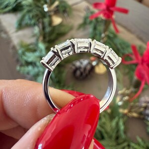 May include: A silver band with five clear, rectangular-shaped gemstones set in a row. The ring is on a hand with red nail polish.