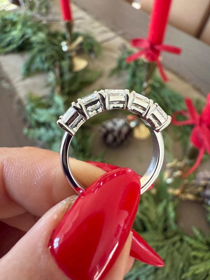 May include: A silver band with five clear, rectangular-shaped gemstones set in a row. The ring is on a hand with red nail polish.