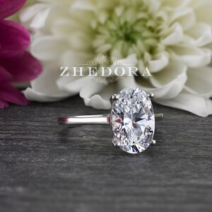 Puede incluir: Un anillo de plata con un gran diamante de forma ovalada. El anillo est&aacute; colocado sobre un fondo de flores blancas y rosas.