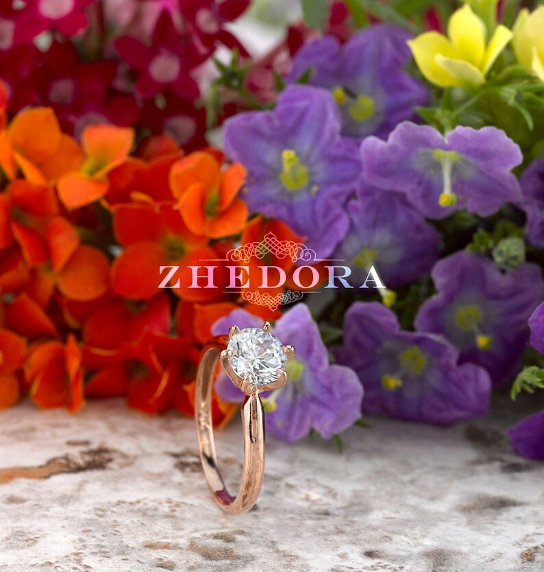 May include: A rose gold engagement ring with a round, clear center stone and a six-prong setting. The ring is surrounded by colorful flowers.