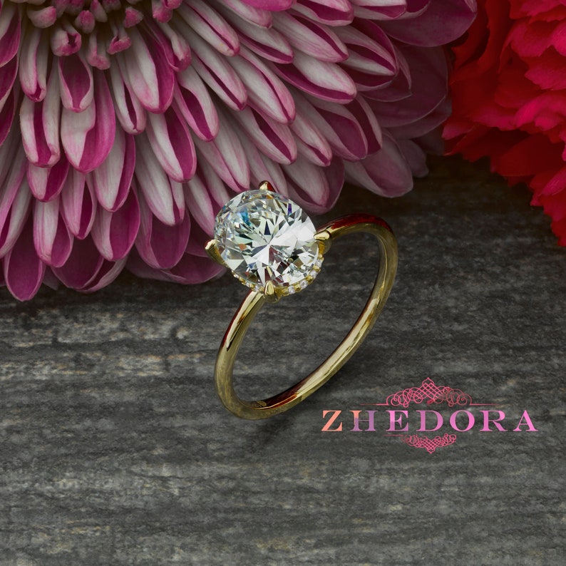 May include: A gold engagement ring with an oval-shaped diamond. The ring is set against a background of pink and red flowers.