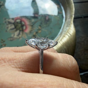 Pu&ograve; includere: Un anello di fidanzamento in argento con un grande diamante trasparente a taglio cuscino. Il diamante &egrave; incastonato su una fascia sottile, tenuto da griffe. Lo sfondo &egrave; sfocato.