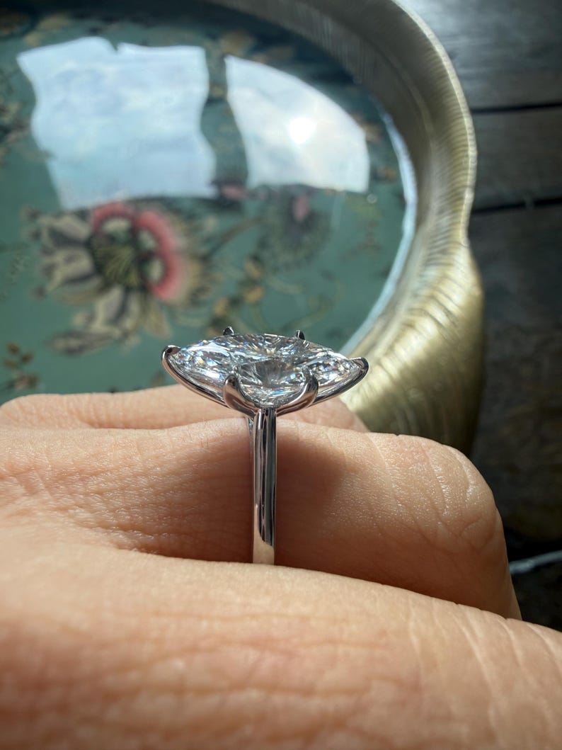 Pu&ograve; includere: Un anello di fidanzamento in argento con un grande diamante trasparente a taglio cuscino. Il diamante &egrave; incastonato su una fascia sottile, tenuto da griffe. Lo sfondo &egrave; sfocato.
