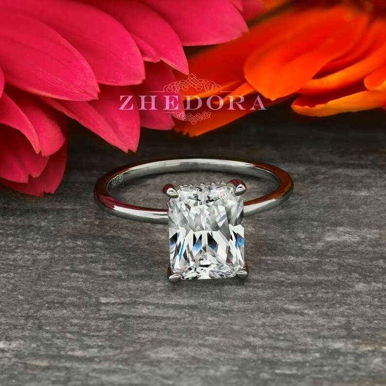 May include: A silver engagement ring with a large, rectangular, clear gemstone. The ring is set on a gray background.