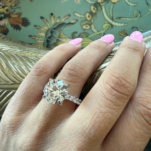 K&ouml;nnte beinhalten: Ein Diamantring mit einem gro&szlig;en, rund geschliffenen Mittelstein und einem mit kleineren Diamanten besetzten Band. Der Ring ist aus Silbermetall gefertigt. Der Hintergrund zeigt ein Blumenmuster.
