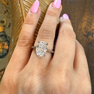 May include: A silver engagement ring with an oval-cut diamond. The ring is set on a slender band and is displayed on a hand with pink nail polish. The background is a dark wood surface.