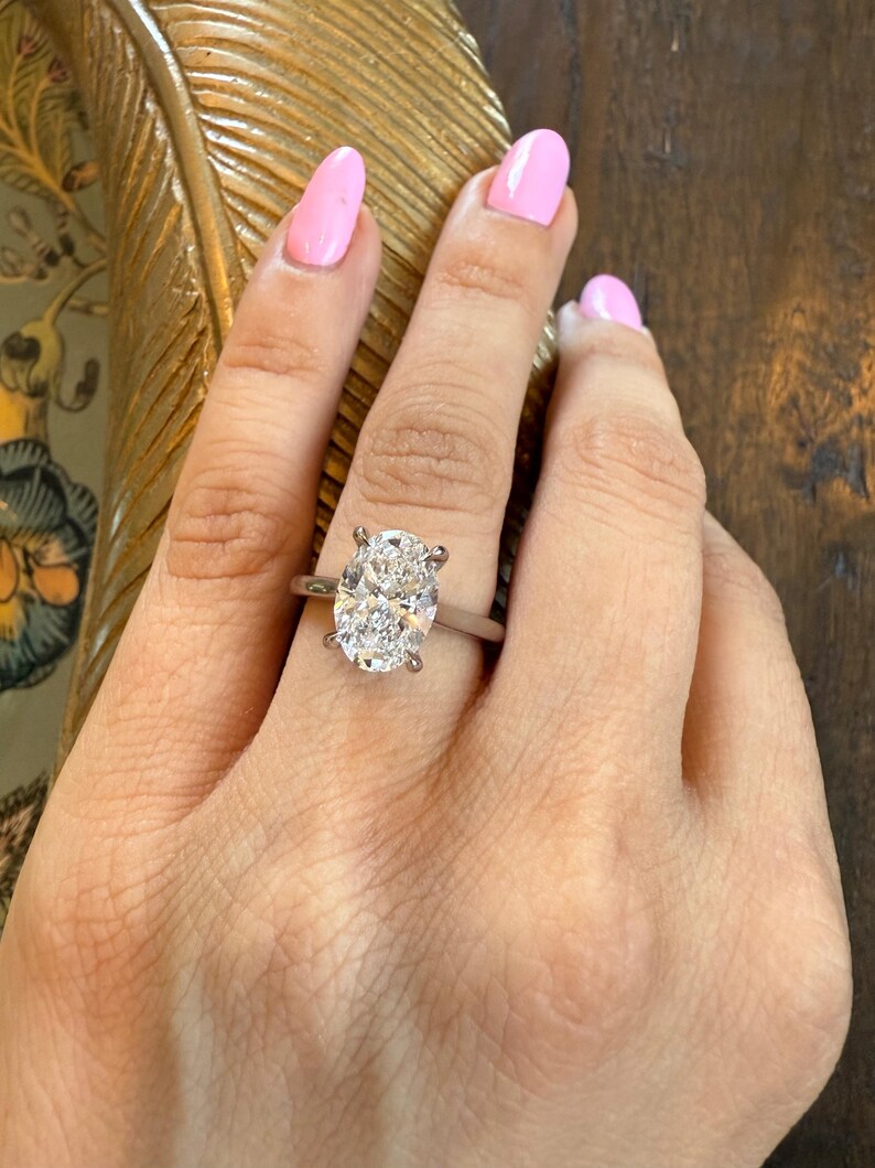 May include: A silver engagement ring with an oval-cut diamond. The ring is set on a slender band and is displayed on a hand with pink nail polish. The background is a dark wood surface.