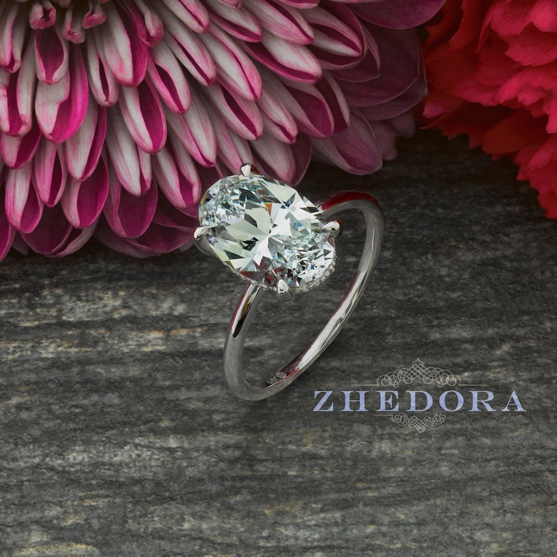 Puede incluir: Un anillo de compromiso de plata con un gran diamante de forma ovalada. El anillo est&aacute; colocado sobre un fondo gris con flores rosas.