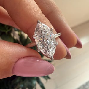 Pu&ograve; includere: Un anello con diamante taglio marquise con fascia argentata. Il grande diamante trasparente &egrave; tenuto da quattro griffe. L'anello &egrave; tenuto su uno sfondo sfocato.