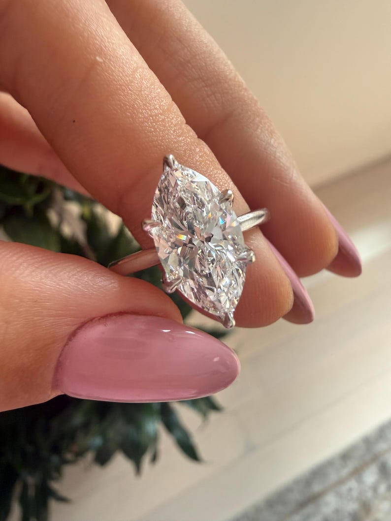 Pu&ograve; includere: Un anello con diamante taglio marquise con fascia argentata. Il grande diamante trasparente &egrave; tenuto da quattro griffe. L'anello &egrave; tenuto su uno sfondo sfocato.