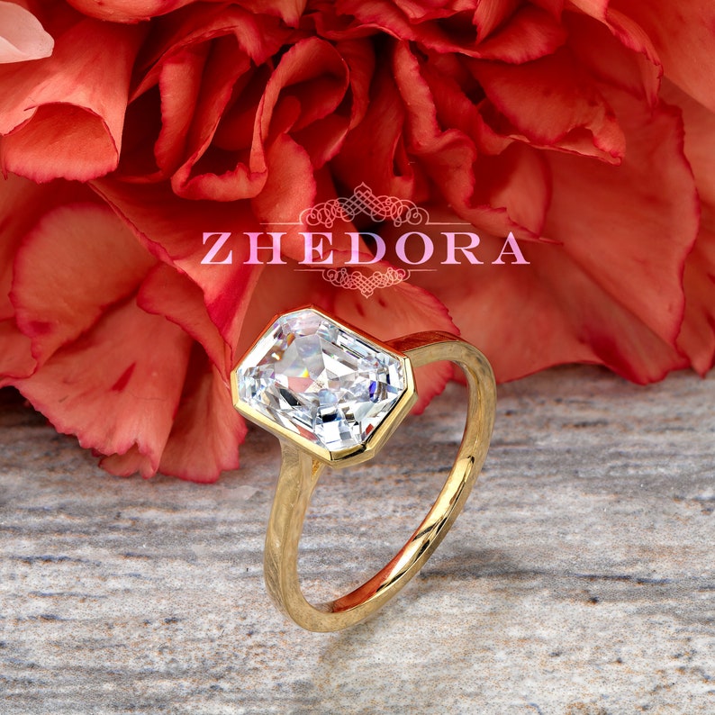May include: A gold engagement ring with a large, clear, rectangular-shaped diamond. The ring is set against a red flower.