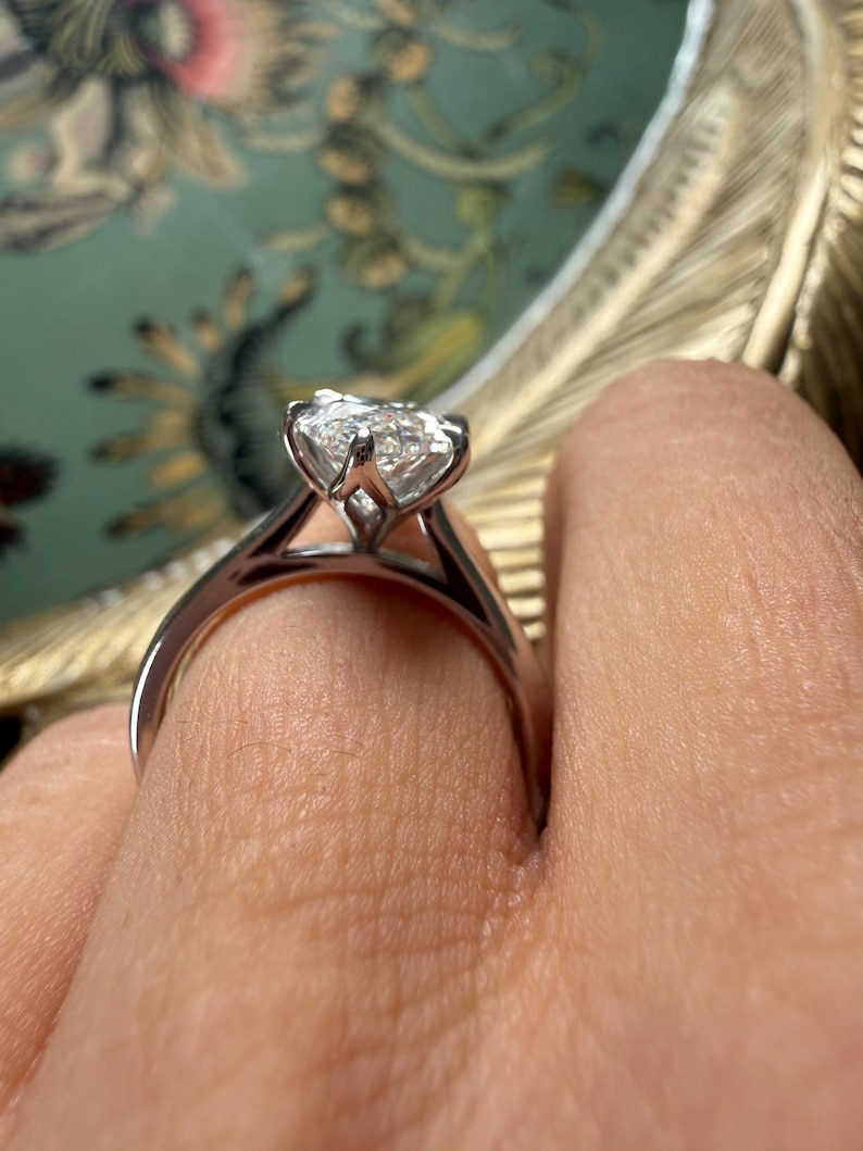 Pu&ograve; includere: Un anello di fidanzamento in argento con un grande diamante trasparente a taglio quadrato. L'anello ha una classica montatura a quattro griffe, che mette in risalto la brillantezza del diamante. La fascia &egrave; liscia e lucida. L'anello &egrave; su un dito.