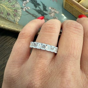 May include: A silver band ring with a row of emerald-cut diamonds. The ring is on a person's finger.