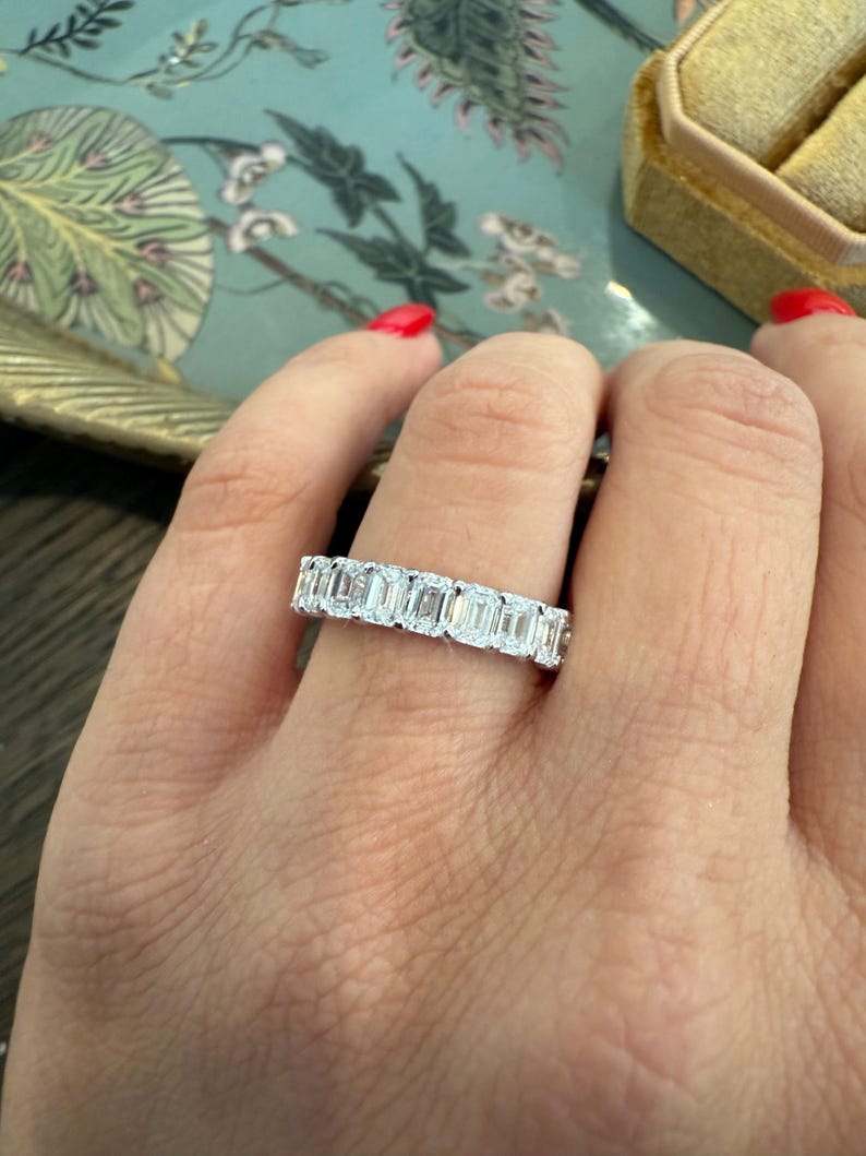 May include: A silver band ring with a row of emerald-cut diamonds. The ring is on a person's finger.