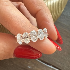 May include: A rose gold ring featuring five oval-cut diamonds. The diamonds are set closely together, creating a continuous sparkle. The ring is held up by a hand with red nail polish, showcasing the ring's brilliance and design.