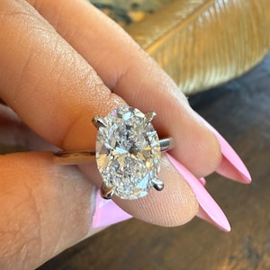 May include: A silver ring with a large, oval-cut diamond. The diamond is clear and sparkling, set with four prongs. The ring is shown against a background of a hand with pink nail polish.