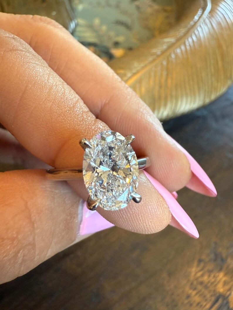 May include: A silver ring with a large, oval-cut diamond. The diamond is clear and sparkling, set with four prongs. The ring is shown against a background of a hand with pink nail polish.