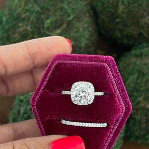 May include: A square-cut diamond engagement ring with a halo setting, and a matching wedding band, both set in silver, displayed in a red velvet ring box.