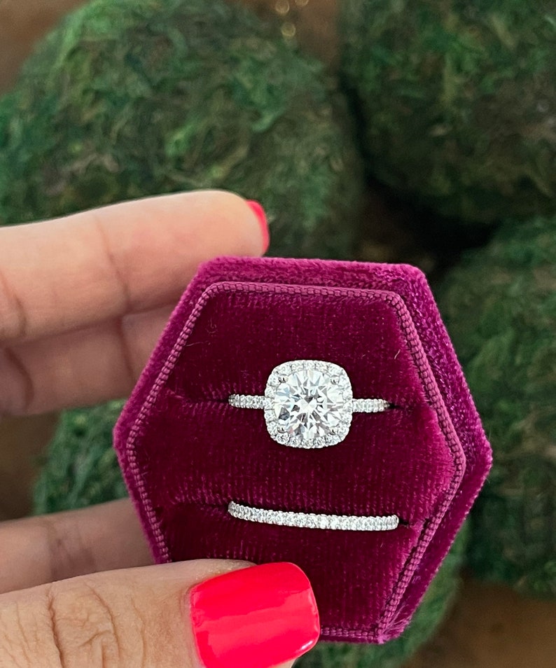 May include: A square-cut diamond engagement ring with a halo setting, and a matching wedding band, both set in silver, displayed in a red velvet ring box.