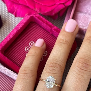 May include: A gold engagement ring with a large oval-shaped diamond. The ring is on a finger with pink nail polish.