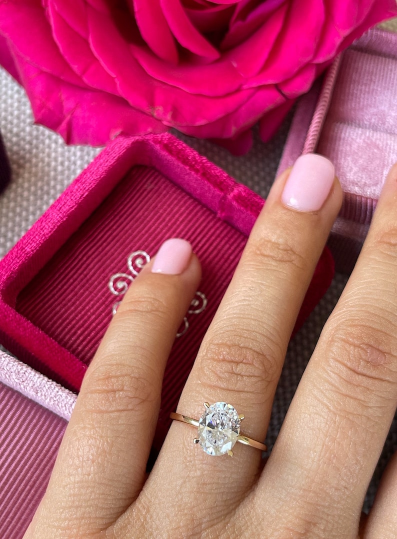 May include: A gold engagement ring with a large oval-shaped diamond. The ring is on a finger with pink nail polish.