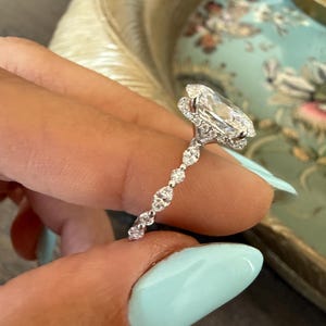 May include: A diamond engagement ring with a large, oval-cut center stone. The ring features a halo of smaller diamonds around the center stone and a delicate band with alternating diamond accents. The ring is held up by a person with light blue nail polish.