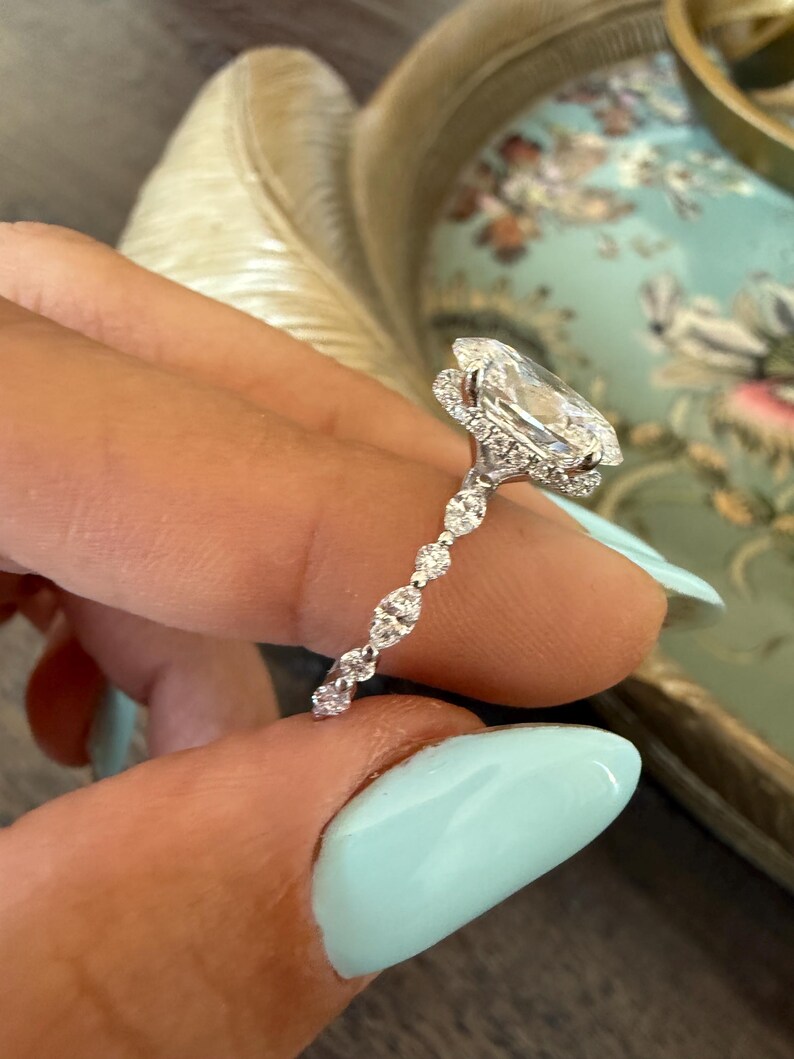May include: A diamond engagement ring with a large, oval-cut center stone. The ring features a halo of smaller diamonds around the center stone and a delicate band with alternating diamond accents. The ring is held up by a person with light blue nail polish.