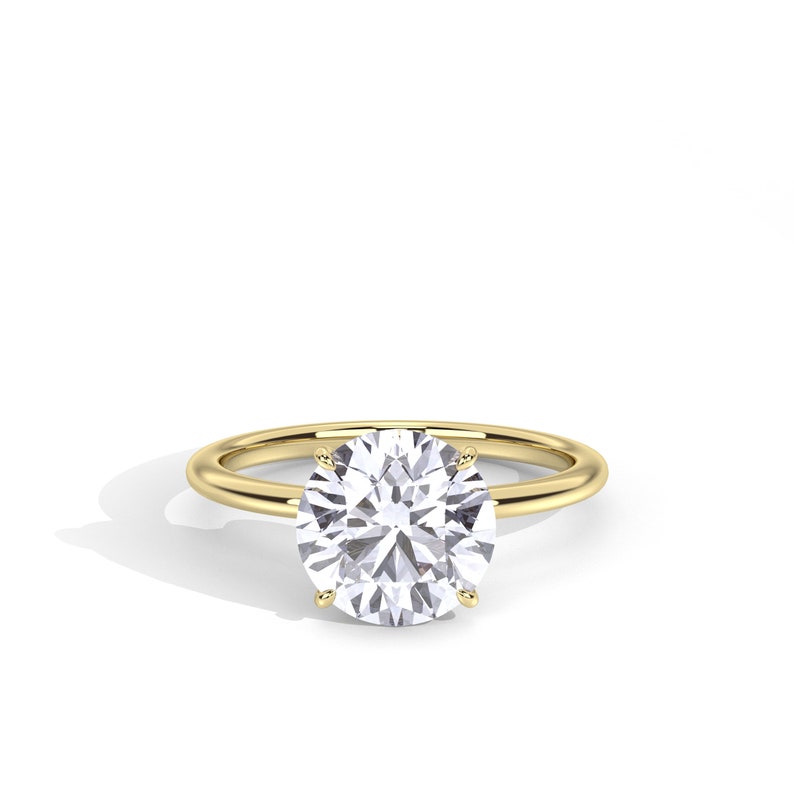 May include: A solitaire engagement ring with a large, round-cut diamond set in a simple, polished gold band. The diamond sparkles brightly against the gold setting, showcasing its clarity and brilliance. The ring is displayed on a white background.