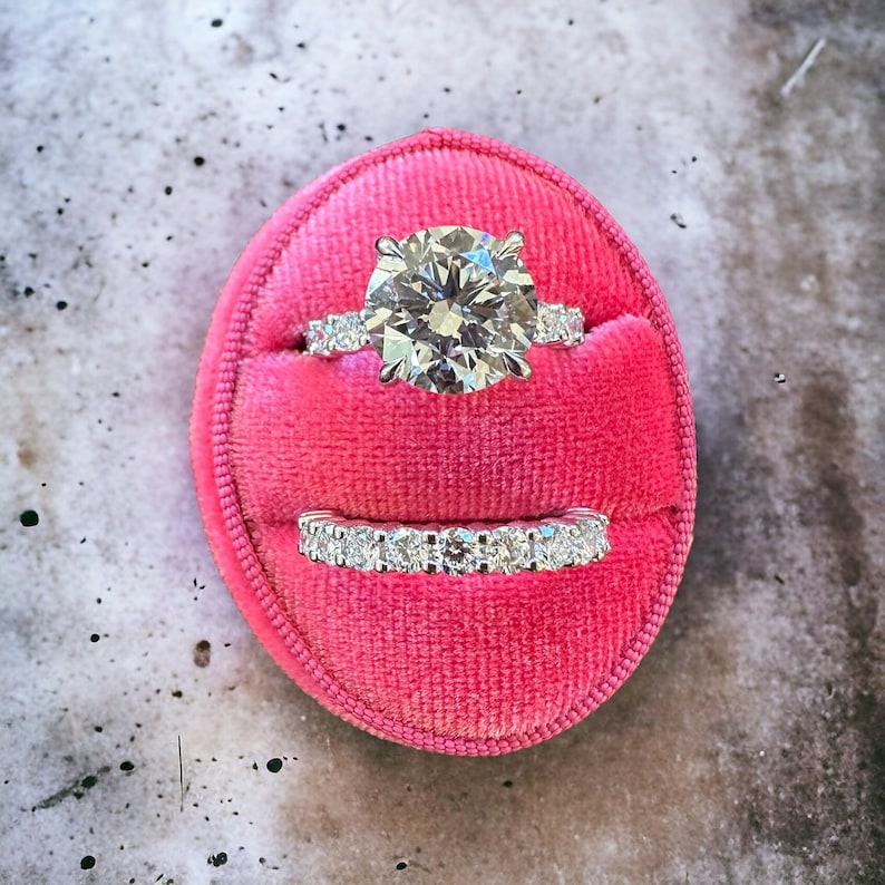 May include: A close-up of a pink velvet ring box with two diamond rings inside. The top ring is a solitaire with a large round diamond, and the bottom ring is a band with smaller diamonds.