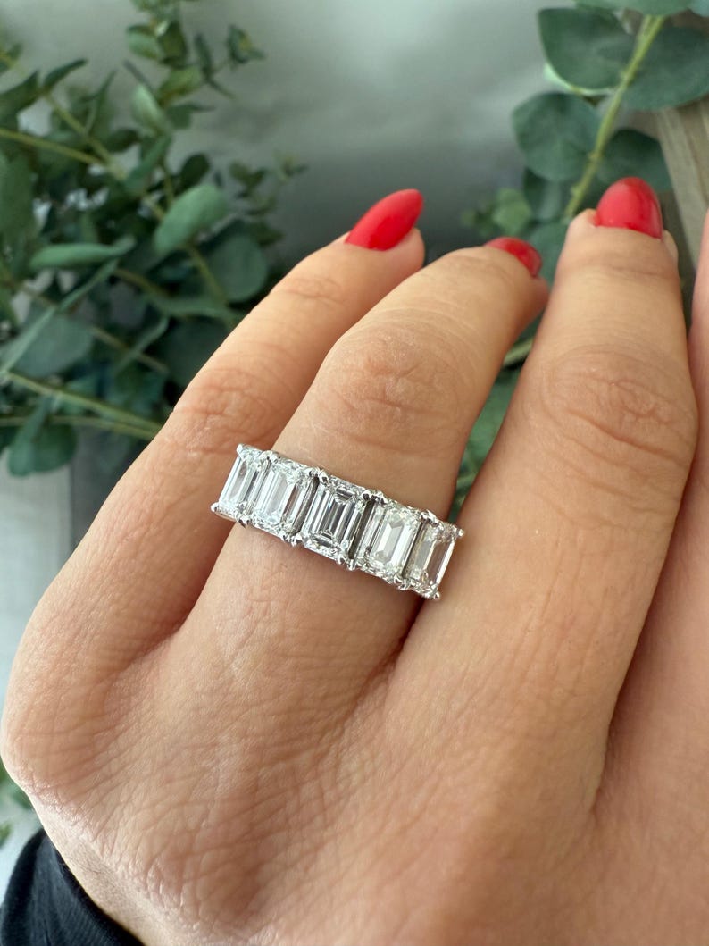 May include: A silver band ring with a row of emerald-cut diamonds. The ring is on a person's finger.
