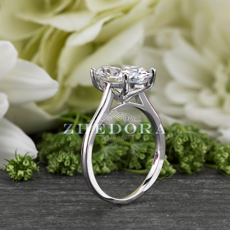 Puede incluir: Un anillo de compromiso de plata con un gran diamante ovalado transparente engastado en garras. El anillo tiene una banda sencilla.