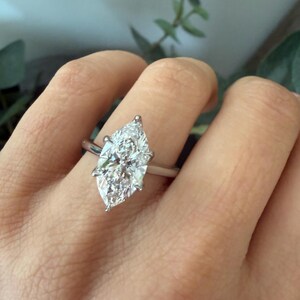 Pu&ograve; includere: Un anello con diamante taglio marquise con una fascia argentata. Il diamante &egrave; incastonato con quattro griffe e brilla intensamente. L'anello &egrave; mostrato su una mano, con uno sfondo sfocato di verde.