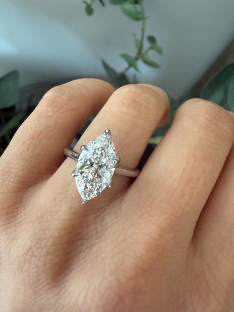 Pu&ograve; includere: Un anello con diamante taglio marquise con una fascia argentata. Il diamante &egrave; incastonato con quattro griffe e brilla intensamente. L'anello &egrave; mostrato su una mano, con uno sfondo sfocato di verde.