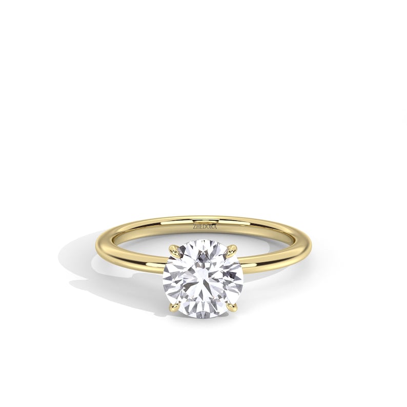 May include: A gold solitaire ring with a large, round, clear gemstone. The ring has a simple, polished band and four prongs holding the gemstone. The brand name "ZIEDORA" is engraved on the band.