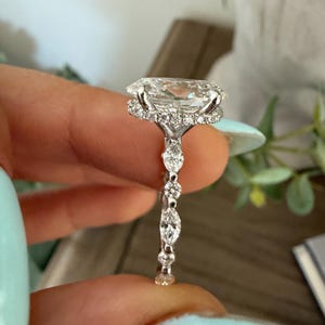 May include: A close-up of a diamond ring with a large, oval-cut center stone. The ring features a halo of smaller diamonds around the center stone and a delicate band with additional diamond accents. The ring is held up against a soft background.