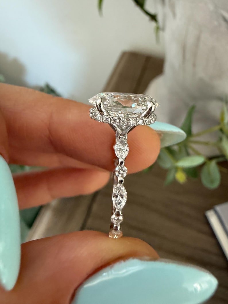 May include: A close-up of a diamond ring with a large, oval-cut center stone. The ring features a halo of smaller diamonds around the center stone and a delicate band with additional diamond accents. The ring is held up against a soft background.