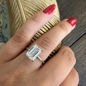 May include: A gold ring with a large, rectangular, clear gemstone. The ring is set on a thin gold band. The gemstone is held in place by four prongs. The background is a wooden surface.