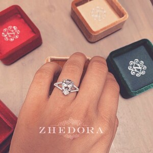 May include: A pear-shaped diamond engagement ring with a split shank and pave setting. The ring is on a hand with a neutral manicure. The ring is in a velvet box with the word "ZHEDORA" printed on the bottom.