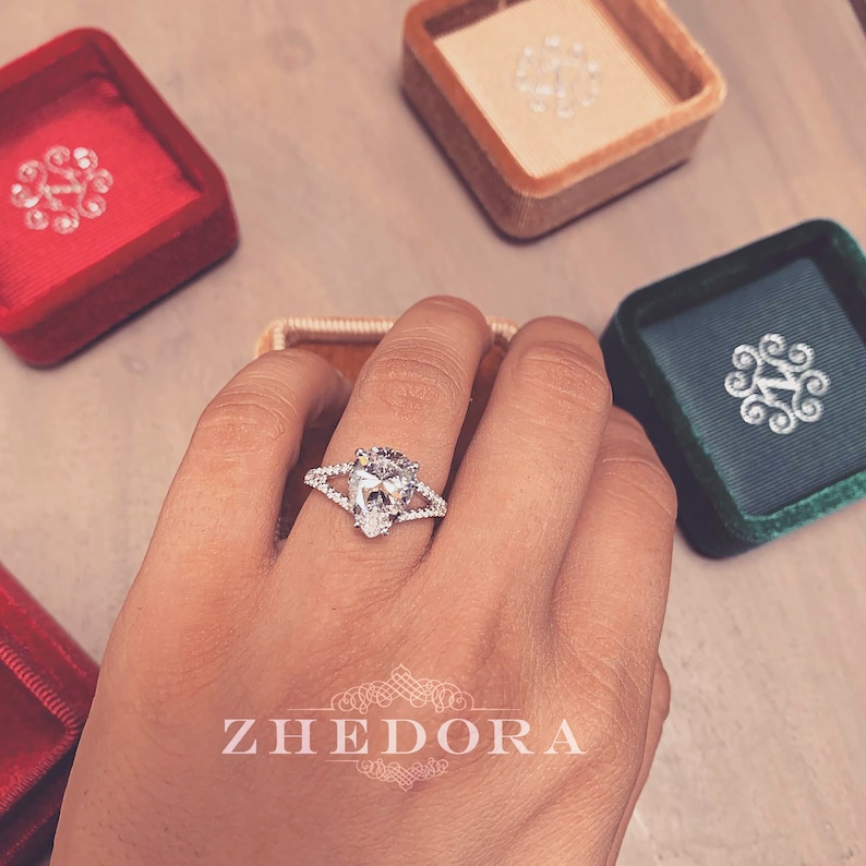 May include: A pear-shaped diamond engagement ring with a split shank and pave setting. The ring is on a hand with a neutral manicure. The ring is in a velvet box with the word "ZHEDORA" printed on the bottom.
