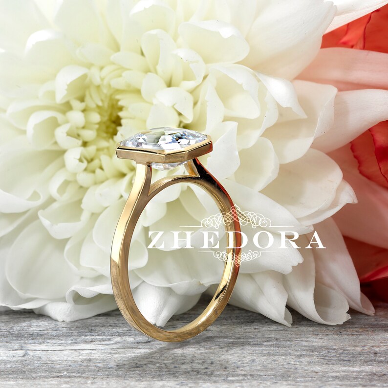 May include: A gold engagement ring with a large, clear, octagonal-shaped diamond. The ring is set on a white background with a white flower.