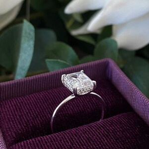 May include: A silver ring with a large, clear, emerald-cut gemstone surrounded by smaller diamonds. The ring is in a purple velvet ring box.