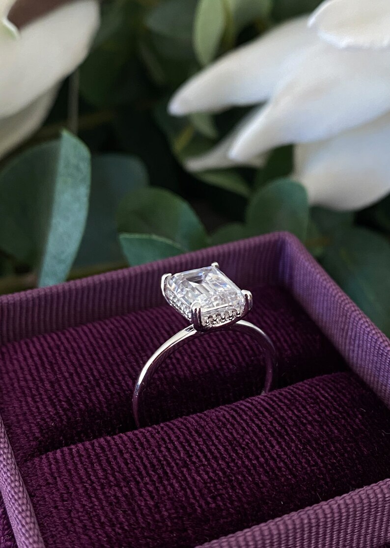 May include: A silver ring with a large, clear, emerald-cut gemstone surrounded by smaller diamonds. The ring is in a purple velvet ring box.