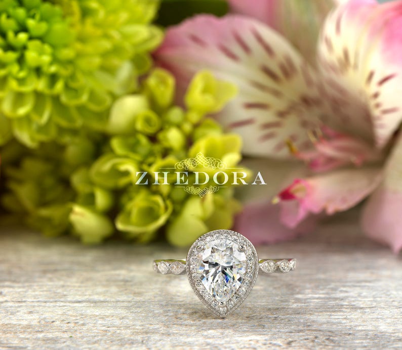 May include: A pear-shaped diamond engagement ring with a halo setting. The ring is set in white gold and features a delicate band with small diamonds.