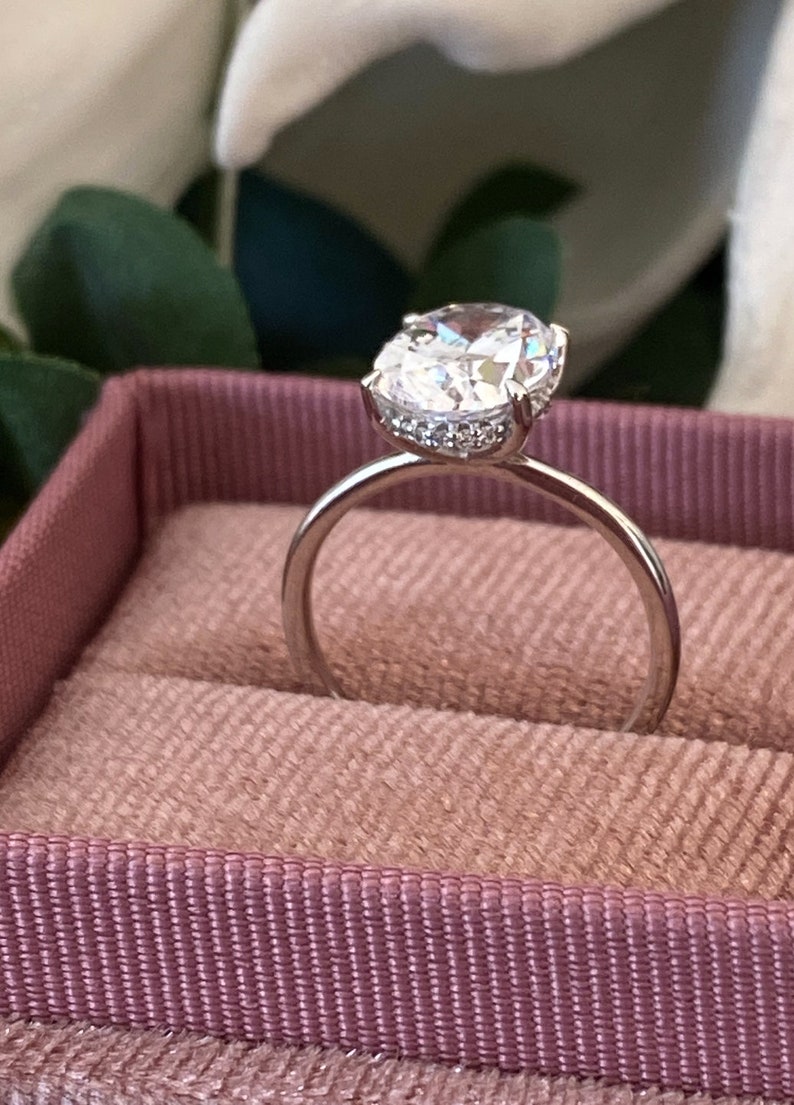 Puede incluir: Un anillo de plata con una gran piedra preciosa ovalada transparente rodeada de piedras preciosas transparentes m&aacute;s peque&ntilde;as. El anillo est&aacute; en una caja de anillo de terciopelo rosa.