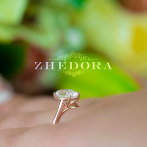 May include: A close-up of a diamond engagement ring with a large oval-shaped center stone set in a delicate prong setting. The ring is on a finger and the background is blurred.