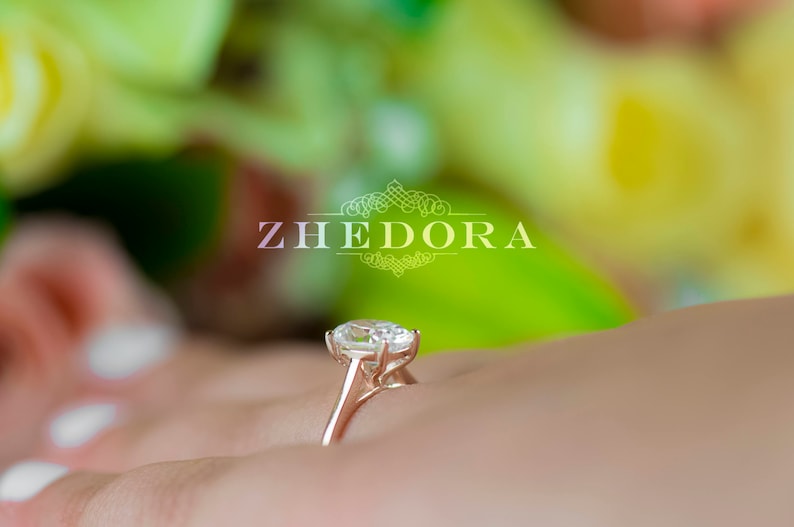 May include: A close-up of a diamond engagement ring with a large oval-shaped center stone set in a delicate prong setting. The ring is on a finger and the background is blurred.